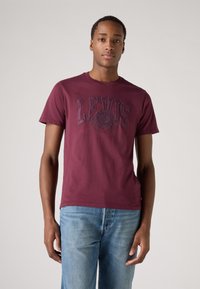 Bordeauxrotes Baumwoll-T-Shirt mit einem erhabenen schwarzen "LEVI'S"-Logo. Kurze Ärmel und ein Rundhalsausschnitt, kombiniert mit hellblauen Jeans.