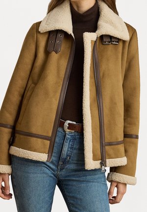 Veste en shearling marron avec doublure et col en polaire crème portée par-dessus un col roulé sombre et un jean bleu avec une ceinture à boucle argentée.