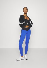 Musta lyhythihainen huppari, jossa on valkoinen logo, yhdistettynä sinisiin leggingsseihin, joissa on mustat sivuviivat ja musta korkea vyötärö. Valkoiset urheilukengät.