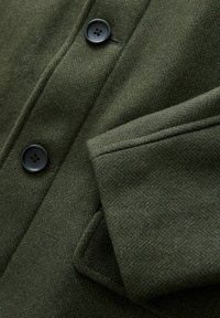 Olivgrüner Wollmantel mit Fischgrätstruktur, ausgestattet mit einem Reverskragen, zwei Knöpfen und einem Detail mit seitlicher Tasche.