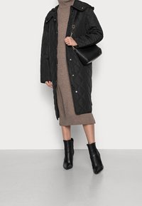 Abrigo largo negro acolchado con capucha, combinado con un vestido de suéter marrón y botines negros. El abrigo cuenta con botones a presión y bolsillos.
