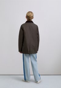 Giacca esterna marrone con colletto, abbinata a jeans a gamba larga celesti con striscia laterale blu. Sneakers bianche completano l'outfit.