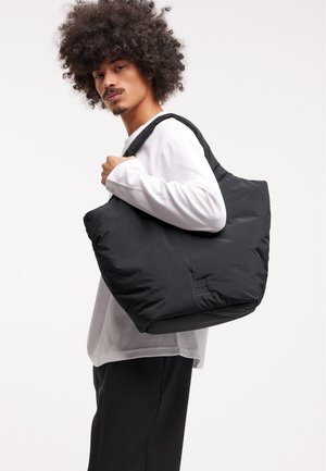 Sort pufet totebag med blød tekstur, der har en stor form og logodetalje. Model iført en lys langærmet top og mørke bukser.