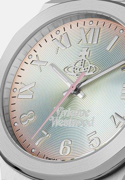 Dichtbijopname van het zilveren horloge van Vivienne Westwood, met Romeinse en Arabische cijfers, een roze secondewijzer en een concentrisch cirkelpatroon.