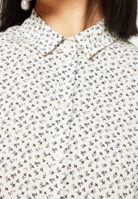Crème blouse met een bloemenpatroon in blauw en zwart, met een knoopsluiting aan de voorkant en een klassieke kraag. Zachte, lichte stof.