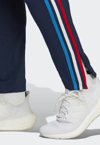 Marineblauwe trainingsbroek met een rode, witte en blauwe zijkantstrips, voorzien van een zijsplit. Gecombineerd met witte sportschoenen en sokken.