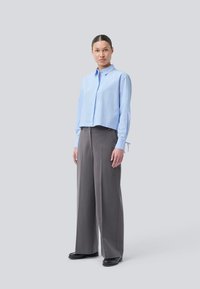 Frau steht in einem hellblauen Langarmshirt und grauen, weit geschnittenen Hosen mit schwarzen Schuhen vor einem schlichten hellen Hintergrund.