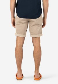 Beige cotton shorts with rolled cuffs, showcasing a relaxed fit. Worn with orange flip-flops, against a white background.