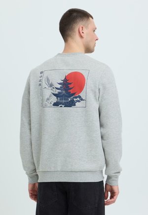 Hombre con sudadera gris claro con un diseño en la espalda que muestra una pagoda azul marino, un sol rojo y una grulla volando, posando frente a un fondo liso.