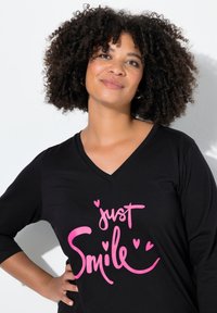 Schwarzes V-Ausschnitt-T-Shirt mit dreiviertelärmeln, auf dem in auffälligem Pink der Text "just Smile" umgeben von kleinen Herzsymbolen steht.
