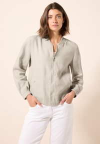 Giacca bomber grigio chiaro con zip frontale, collo arricciato e polsini elasticizzati. Presenta due tasche laterali. Abbinata a pantaloni bianchi.