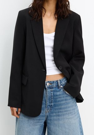 Femme portant un blazer noir sur un débardeur court blanc et un jean bleu taille haute, avec une main dans la poche.