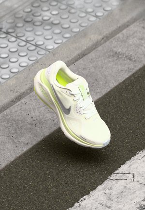 Biały i neonowo-żółty but do biegania Nike zawieszony w powietrzu nad strukturalnym krawężnikiem i asfaltową nawierzchnią drogi.
