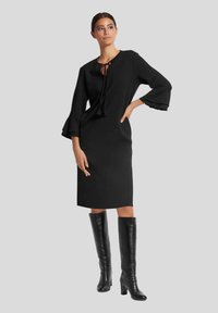 Schwarzes knielanges Kleid mit langen, ausgestellten Ärmeln und einer Schleifenhalsdetail, kombiniert mit knietiefen schwarzen Lederstiefeln. Glatte Stofftextur.