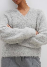 Helles graues, flauschiges Sweater mit V-Ausschnitt und breiten, weichen Ärmeln. Strukturierte Oberfläche mit einem gemütlichen Aussehen, kombiniert mit maßgeschneiderten grauen Hosen.