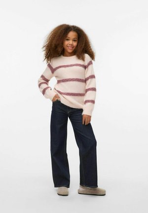 Giovane con capelli ricci che indossa un maglione a righe, jeans larghi scuri e pantofole beige, in piedi con una mano in tasca.
