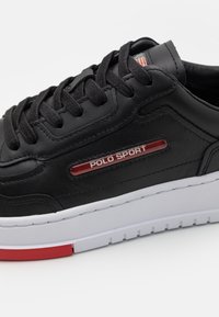 Baskets en cuir noir avec lacets noirs, accents rouges et marque "POLO SPORT". Elles disposent d'une semelle blanche avec des détails rouges et une surface texturée.