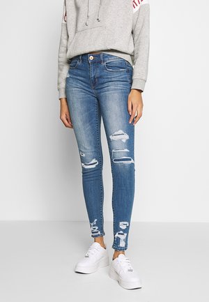 Osoba ubrana w podarte, niebieskie, dopasowane jeansy, szarą bluzę z kapturem i białe trampki, stojąca na tle jednolitego, białego tła.