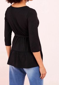 Žena v černém, dlouhém peplum topu s detailem pásku a světle modrými džíny, viděná zezadu na světlém pozadí.