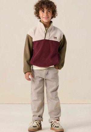 Fleece pullover in cream, bordeaux en olijfgroen, met een hoge kraag en knoopsluiting. Gecombineerd met lichte jeans en gemusterde sneakers.