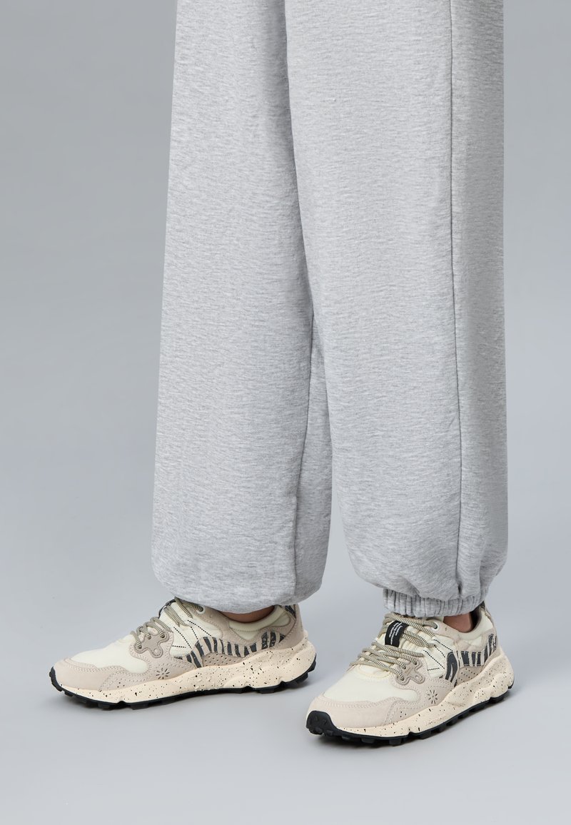 Person trägt hellgraue Jogginghose und beige Sneaker mit schwarzen Zebramustern und gesprenkelten Sohlen auf grauem Boden.