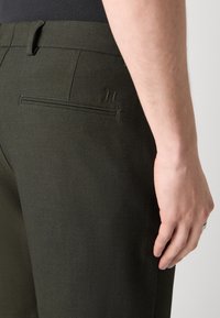 Gros plan d'une personne portant un pantalon vert foncé sur mesure avec une poche passepoilée arrière et des passants de ceinture, main reposant sur le côté.