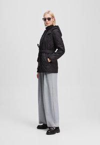Veste noire matelassée avec capuche et taille ceinturée, associée à un pantalon large gris clair et des chaussures noires imposantes. Offrant une silhouette moderne.