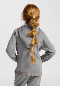 Grå hættetrøje med en tekstureret finish og en snøre i hætten, vist bagfra. Håret er stylet i en fletning med en elastik.