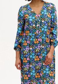 Robe florale à manches longues avec un décolleté en V. Présente des fleurs multicolores sur un fond bleu foncé, une taille froncée et des accents de boutons.
