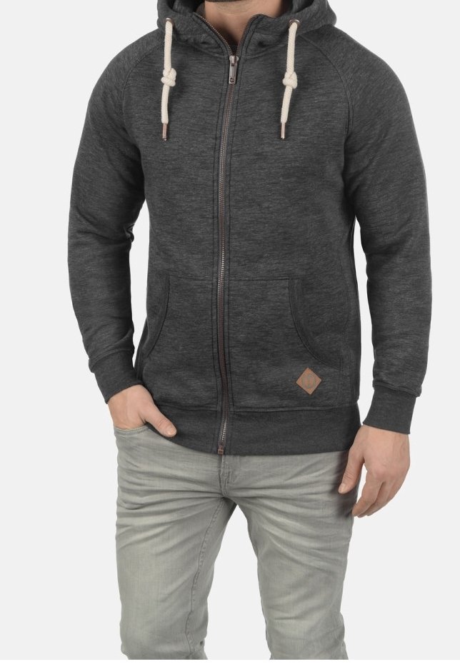 Homme portant un sweat à capuche gris foncé zippé avec des cordons blancs et un jean gris clair, debout avec une main dans la poche.
