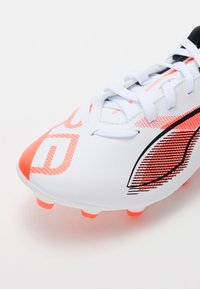 Hvit fotballsko med oransje aksenter, teksturert overdel og spiss tå. Har snøring og rosa grepstuds på sålen.
