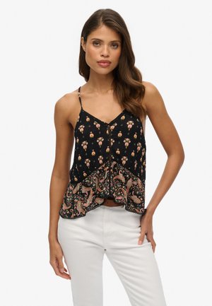 Kobieta w czarnym, bezrękawnikowym topie z kwiatowymi i paisley wzorami, w białych spodniach, stojąca na tle jednolitego białego tła.