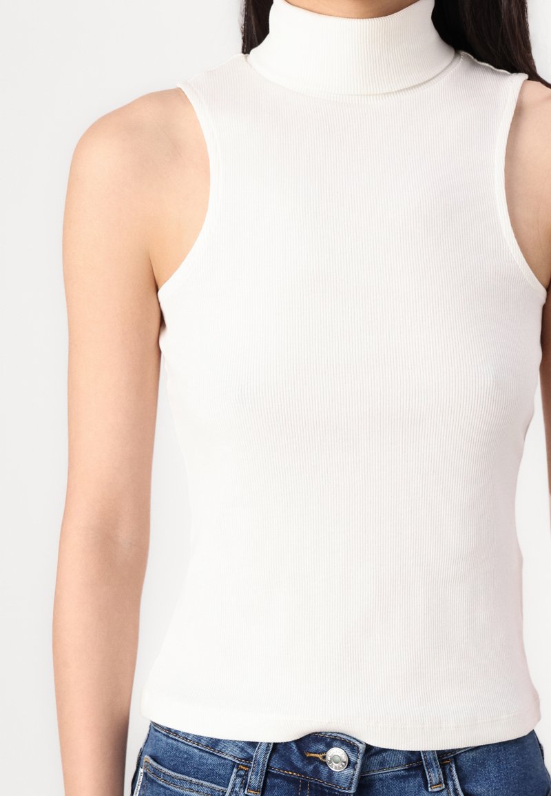 Top bianco a costine senza maniche con un design aderente e una texture liscia, abbinato a jeans in denim blu con un bottone metallico.