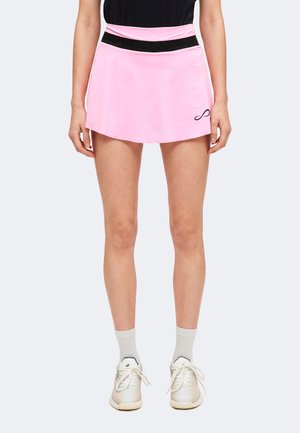 Vrouw met een roze tennisrokje met een zwarte tailleband, witte sokken en witte sneakers, staand tegen een effen achtergrond.