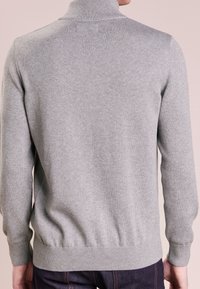 Mann trägt einen hellgrauen Langarmpullover und dunkle Jeans, von hinten vor einem schlichten Hintergrund gezeigt.