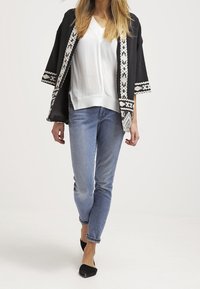 Kvinde iført en sort-hvid mønstret åben jakke, hvid V-hals bluse, lyseblå jeans og sorte spidse flade sko gående fremad.