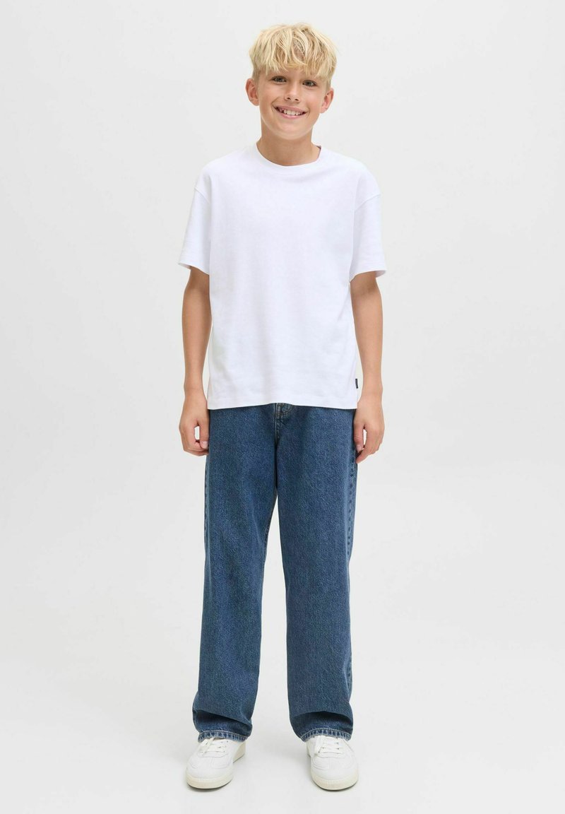 T-shirt blanc à manches courtes associé à un jean en denim bleu ample et des baskets blanches, posé sur un fond clair et uni.