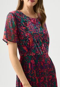 Blumenmuster-Kleid mit grünem Grund und rosa sowie lila Akzenten, leichtem Stoff, plissiertem Rock und kurzen, lockeren Ärmeln.