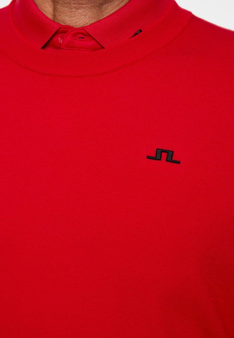 Roter Pullover über einem roten Hemd mit Kragen, mit einem kleinen schwarzen geometrischen Logo auf der linken Brust.