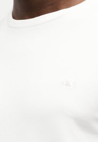 Κοντινό πλάνο ενός ατόμου που φοράει ένα απλό λευκό t-shirt με στρογγυλή λαιμόκοψη και μικρό ανάγλυφο λογότυπο "CK" στο στήθος.