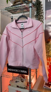 Ανοιχτό ροζ μπουφάν Nike με φερμουάρ και κόκκινες γραμμές στο μπροστινό μέρος, κρεμασμένο σε μαύρη κρεμάστρα σε μεταλλική ραφιέρα με παπούτσια και φυτά πίσω.
