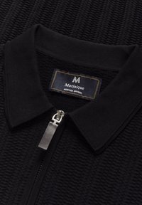 Schwarzes Strick-Poloshirt mit strukturiertem Muster, silbernem Reißverschluss und kontrastierendem Kragen. Mit einem Label "Matinique Heritage Apparel."