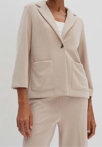 Beige lehký blazer s jedním knoflíkem, tříčtvrtečními rukávy, dvěma náprsními kapsami a hladkou texturou látky. Střih na míru.