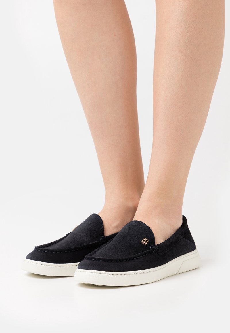 Chaussures à enfiler en daim noir avec une semelle blanche, ornées de coutures décoratives et d'un petit logo doré sur le côté. Texture lisse.