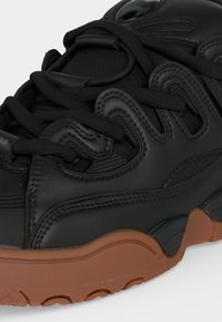 Gros plan d'une basket noire avec une tige en cuir texturé ondulé et une semelle en caoutchouc marron, montrant des coutures détaillées et de gros lacets noirs.