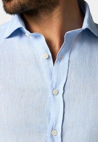 Chemise en lin bleu clair avec un col classique, petite patte de boutonnage et texture naturelle, comportant des bords légèrement effilochés.