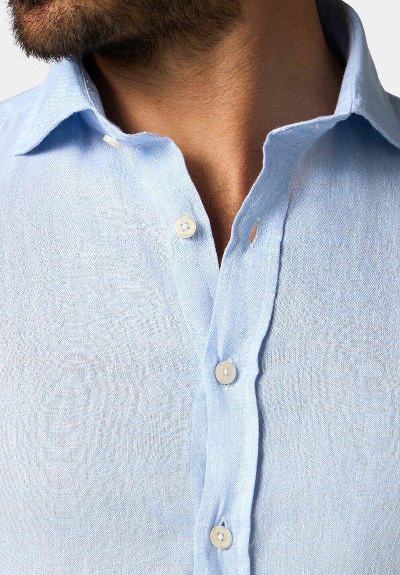 Chemise en lin bleu clair avec un col classique, petite patte de boutonnage et texture naturelle, comportant des bords légèrement effilochés.
