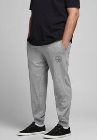 Pantalones deportivos de algodón gris con bolsillos laterales y un parche redondo del logotipo en el muslo. Combinados con zapatillas negras. Diseño minimalista.