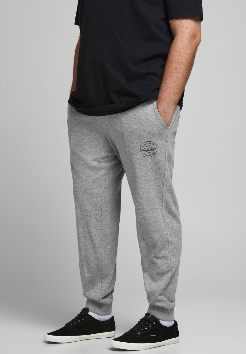 Pantalones deportivos de algodón gris con bolsillos laterales y un parche redondo del logotipo en el muslo. Combinados con zapatillas negras. Diseño minimalista.