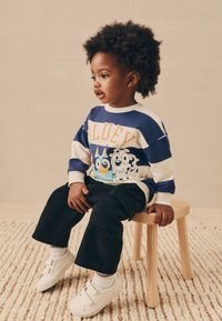 Felpa a righe blu e bianche con personaggi dei cartoni animati, pantaloni neri e sneaker bianche, seduto su uno sgabello di legno chiaro.